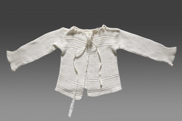 Kaftanik dziecięcy w kolorze écru. Ścieg układa się w geometryczny wzór. W centralnej części obiektu widoczny wzór kwadratów układających się w równoległe rzędy biegnące z góry do dołu. Na piesi oraz w dolnej części (około 1/3 wysokości od dolnej krawędzi) wzór układa się w dwa równoległe poziome pasy. Pomiędzy nimi ścieg jest równomierny (bez podziału na rzędy). Dolna krawędź wykończona falbanką. Kaftanik wiązany pod szyją na taśmę w tym samym kolorze. Rękawy zakończone ściągaczem.