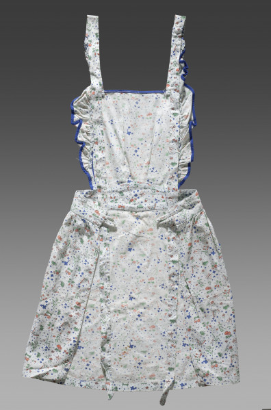 Fartuszek dziecięcy wykonany z tkaniny w kolorze écru z ozdobnym kolorowym nadrukiem – drobne wizerunki kwiatów, liści, motyli, ważek i grzybów (muchomorów). Nadruk w kolorach: zielonym, niebieskim i pomarańczowym. Fartuch składa się z prostokątnego fragmentu materiału obszytego po bokach falbanką wykończoną niebieską lamówką (karczek/część piersiowa), paska oraz spódnicy. Karczek, pasek oraz spódnica zostały ze sobą zszyte. Do górnych krawędzi karczka przyszyte zostały długie pasy tkaniny (umożliwiające jego zawiązanie na szyi). Pasek zakończony jest na obu końcach pętelkami wykonanymi z tej samej tkaniny (miejsce na guzik lub tasiemkę). Do spódnicy zostały przyszyte kieszenie obszyte dodatkowo ozdobną niebieską lamówką.