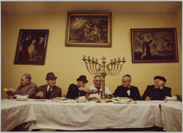 fotografia - Zdjęcie przedstawia grupę sześciu mężczyzn siedzących za stołem podczas wieczerzy sederowej. Mężczyźni siedzą po jednej stronie stołu, pod ścianą. Po środku stołu widoczny ozdobny ośmioramienny świecznik (miejsca na świece są puste). Stół jest nakryty białym obrusem i zastawiony potrawami. Mężczyźni są ubrani w garnitury oraz nakrycia głowy (jarmułki lub kapelusze). Trzeci mężczyzna od lewej nalewa wino do kieliszka drugiego mężczyzny od prawej strony. W tle, na ścianie widoczne trzy obrazy w ozdobnych ramach.