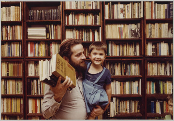 fotografia - Zdjęcie przedstawia mężczyznę z małą dziewczynka (córeczką) na ręku stojącego na tle regału z książkami (wypełniającego całe tło). Mężczyzna ma na sobie jasną koszulę i jarmułkę na głowie. Dziewczynka jest ubrana w niebieską sukienkę oraz ciemną koszulkę. Twarz mężczyzny jest zwrócona w stronę dziecka. Dziewczyna patrzy w obiektyw i się uśmiecha. W drugim ręku mężczyzna trzyma kilka książek.