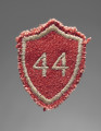 Tarcza szkolna w formie naszywki wykonana z tkaniny wełnianej w kolorze czerwonym. Czworoboczna - z trzema rogami na górze i jednym na dole, każda krawędź zaokrąglona: górne do wewnątrz (do dołu), boczne na zewnątrz. Krawędzie obszyte nicią w kolorze żółtobrązowym. W centralnym polu wszyty maszynowo nicią w tym samym kolorze numer ,,44’’ (nr szkoły). Po stronie verso płócienna podkładka w kolorze białym.