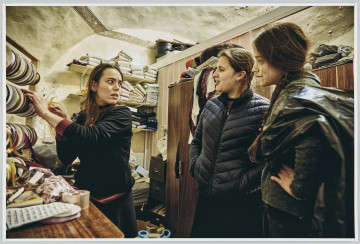 scena rodzajowa - Poziomy, barwny kadr fotograficzny przedstawiający 3 nastolatki stojące i rozmawiające w pomieszczeniu. Jedna z nich, po lewej stronie kadru, sięga po wstążkę na ze stojaka pełnego rolek ze wstążkami i odwraca się w stronę dwóch pozostałych dziewcząt. Pomieszczenie jest niewielkie, zabałaganione i zaniedbane (purchle na suficie), pełne jest ubrań i elementów krawieckich oraz pasmanteryjnych.