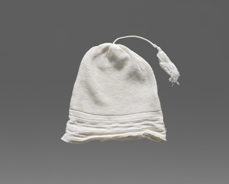 Czapeczka dziecięca w kolorze białym/écru. W dolnej części (około 1/3 wysokości od dolnej krawędzi) widoczny ozdobny geometryczny ścieg w formie szachownicy. W górnej części (po środku) przyszyty został sznurek zakończony frędzlem.