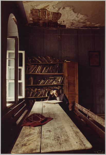 fotografia - Zdjęcie przedstawia starszego mężczyznę siedzącego przy drewnianym stole. Zajmuje miejsce na końcu stołu, w głębi fotografii. Siedzi na jednej z drewnianych ław z oparciem, które są ustawione po obu jego stronach. Mężczyzna opiera się łokciem o stół, a głowę podpiera o dłoń. Przed nim leży otwarta księga, na której kartach mężczyzna skupia wzrok. Na stole leży także bordowa tkanina ozdobiona haftem sześcioramiennej gwiazdy (gwiazdy Dawida). Mężczyzna ma na sobie ciemny garnitur w drobne prążki oraz jarmułkę na głowie. Za nim (w tle) widoczny regał uginający się pod ciężarem licznych ksiąg. Po lewej stronie kadru widoczne otwarte okno. Na górze kadru sufit z widocznym ubytkiem tynku (zawilgocenie, spod tynku wyzierają deski).