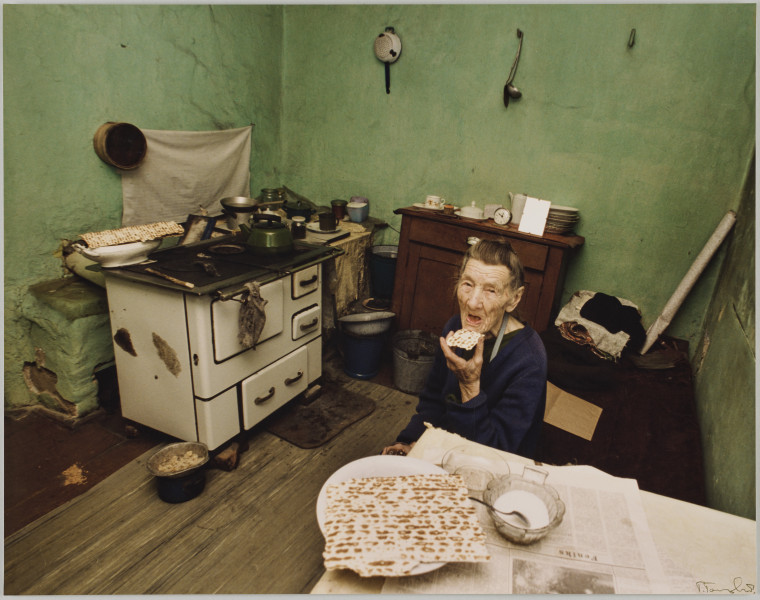 fotografia - Zdjęcie przedstawia starszą kobietę siedzącą przy stole w kuchni. Kobieta unosi do ust kawałek macy. Na stole leży rozłożona gazeta, talerz z płatem macy oraz cukiernica i pusty talerz (stojący przed nią). Po lewej stronie kadru widać białą kuchenkę (westfalkę), a na niej naczynia oraz białą wazę. Na wazie leży kolejny płat macy. Za kuchenką znajdują się dwie szafki, na nich ceramiczne naczynia. Na podłodze, pod kuchenką znajduje się blaszana miska z resztkami.