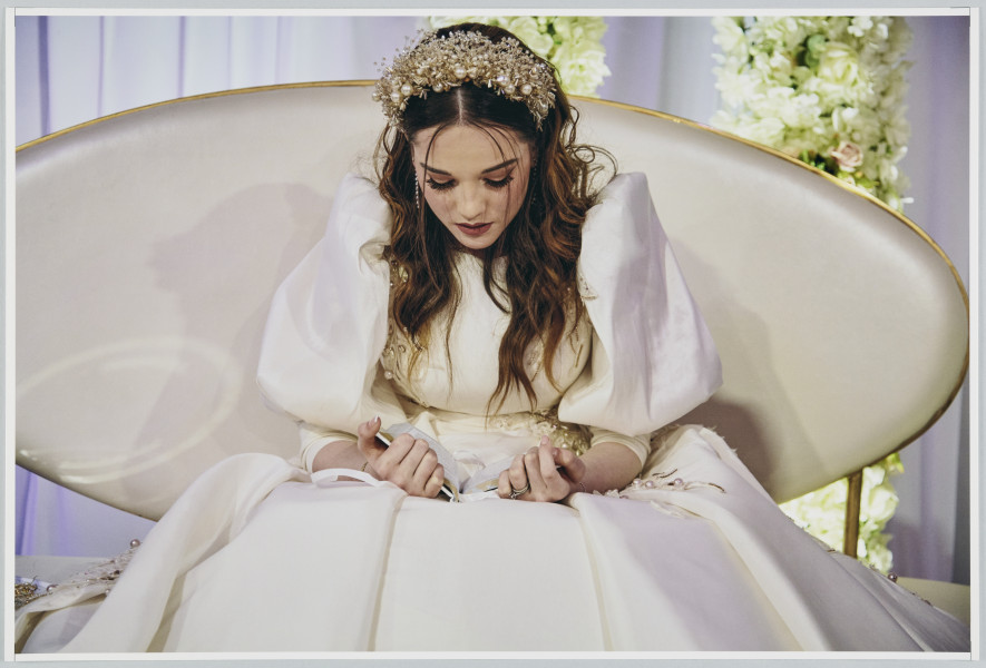 scena rodzajowa - Poziomy, barwny kadr fotograficzny przedstawiający portret kobiety (panny młodej) ubranej w białą suknię z połyskliwej tkaniny, z boku gorsetu i na górnym fragmencie spódnicy obszytą haftem oraz perełkami. Na twarzy ma makijaż, a rozpuszczonych włosach - ozdobny stroiku. Kobieta siedzi na ławce tapicerowanej na biało (skórą?) o owalnym oparciu na ze złotą obwódką stojącej na tle białej udrapowanej zasłony i dwóch słupów udekorowanych białymi kwiatami. Na kolanach trzyma książeczkę, która pochylona czyta.