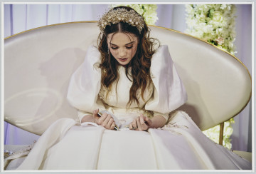 scena rodzajowa - Poziomy, barwny kadr fotograficzny przedstawiający portret kobiety (panny młodej) ubranej w białą suknię z połyskliwej tkaniny, z boku gorsetu i na górnym fragmencie spódnicy obszytą haftem oraz perełkami. Na twarzy ma makijaż, a rozpuszczonych włosach - ozdobny stroiku. Kobieta siedzi na ławce tapicerowanej na biało (skórą?) o owalnym oparciu na ze złotą obwódką stojącej na tle białej udrapowanej zasłony i dwóch słupów udekorowanych białymi kwiatami. Na kolanach trzyma książeczkę, która pochylona czyta.