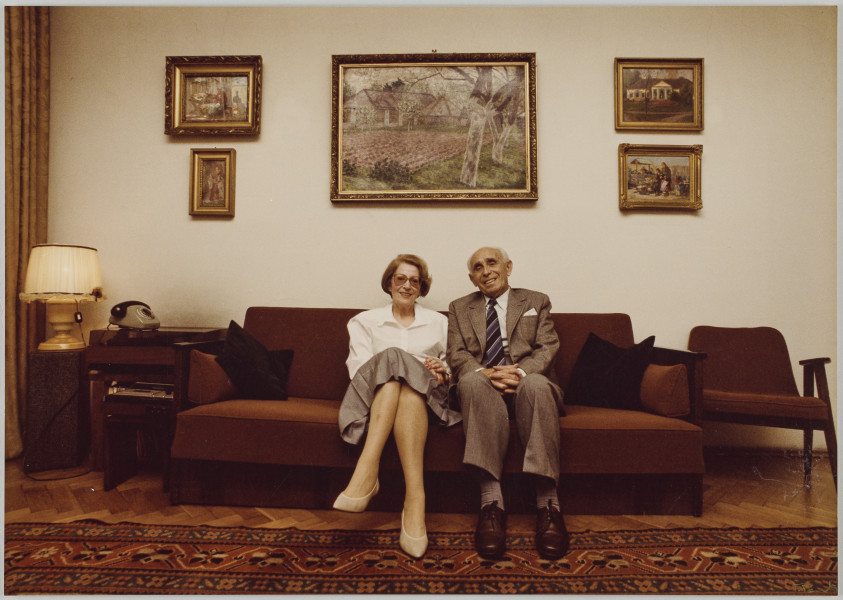 fotografia - Zdjęcie przedstawia parę starszych osób siedzących na bordowej kanapie. Mężczyzna (siedzący po prawej stronie) ma na sobie szary garnitur, białą koszulę i krawat. Jest gładko ogolony. Kobieta ma na sobie szarą spódnicę, białą bluzkę i okulary. Zakłada nogę na nogę. Oboje uśmiechają się do obiektywu. Przed kanapą widoczny dywan. Po lewej stronie kanapy szafka, na niej telefon stacjonarny, a dalej ozdobna lampa. Po prawej stronie kanapy widoczny tapicerowany (również bordowy) fotel z drewnianymi podłokietnikami. Za kanapą, na jasnej ścianie wiszą obrazy. 