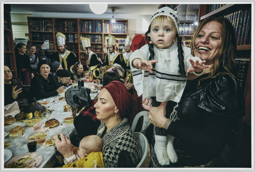 scena rodzajowa - Poziomy, barwny kadr fotograficzny przedstawiający ucztującą grupę kobiet, mężczyzn i dzieci we wnętrzu. Na pierwszym planie, po prawej stronie, widać dwie kobiety z niemowlętami – jedna z nich siedzi przy stole trzymając dziecko na kolanach, druga stoi, śmiejąc się prezentuje na rękach niemowlę w kostiumie przebierańców. Jej sylwetka zajmuje całą prawą krawędź kadru. Od lewego narożnika, pod kątem, w głąb sceny biegnie długi stół zastawiony jedzeniem i piciem. Po obu jego stronach siedzą kobiety i dzieci, za nim widać postaci 4 mężczyzn o fryzurach z charakterystycznymi długimi, zakręconymi pasmami nad uszami (pejsami) poprzebieranych w jednakowe kostiumy. W głębi, po lewej stronie stoi kilka kobiet.