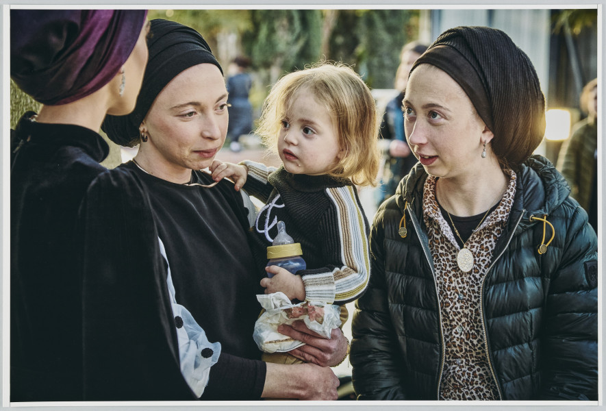 scena rodzajowa - Poziomy, barwny kadr fotograficzny przedstawiający 3 młode kobiety stojące na ulicy. Jedna z nich stojąca tyłem do widza, a przodem do pozostałych, ubrana jest w czarny strój, na głowie ma ciemnofioletową chustę zawiązaną wokół głowy. Kobieta stojąca w centrum kadru ubrana jest w czarny sweter o białych rękawach w czarne kropki, na głowie ma czarną chustę zawiązaną także wokół głowy. Na lewym ręku trzyma dziecko o jasnych włosach do ramion, które w lewej ręce trzyma butelkę ze smoczkiem. Patrzy z zamyśleniem przed siebie. Trzecia uczestniczka sceny stoi po prawej stronie ubrana jest w czarna koszulkę, bluzkę z wzorem panterki oraz rozpiętą czarna kurtkę. Na głowie nosi czarna chustę zawiązana podobnie jak poprzednie. Twarz ma zwróconą w stronę kobiety stojącej tyłem, mimika twarzy wskazuje na to, że mówi.