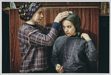 scena rodzajowa - Poziomy, barwny kadr fotograficzny przedstawiający dwie kobiety ubrane w swetry w stonowanych kolorach oraz chusty zawiązane na głowach, stojące w centralnej części kadru na tle szafy na ubrania, we wnętrzu mieszkalnym. Jedna z kobiet poprawia drugiej chustę.