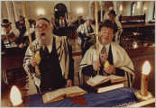 fotografia - Zdjęcie przedstawia wnętrze synagogi oraz modlących się mężczyzn. Na pierwszym planie widoczni są dwaj mężczyźni. Stoją przy pulpicie, na którym leżą otwarte księgi. Obaj mają na sobie garnitury, białe koszule i krawaty. Ich ramiona są okryte tałesami, na głowach jarmułki. Obaj trzymają w rękach cytrynę (lub etrog) oraz lulaw (wiązankę roślin wykorzystywaną w modlitwie wraz z etrogiem siódmego dnia Sukot, w dzień Hoszana Raba). Za nimi widoczne wnętrze synagogi, a w nimi pozostali modlący się mężczyźni. Wielu z nich ma ramionach okryte tałesem. 