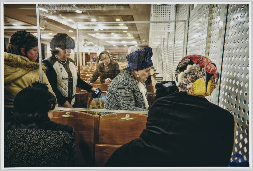 scena rodzajowa - Poziomy, barwny kadr fotograficzny przedstawiający kilka kobiet stojących w dużym wnętrzu wypełnionym rzędami krzeseł i patrzących przez prześwity ażurowej ściany znajdującej się po prawej stronie, wydzielające pomieszczenie.