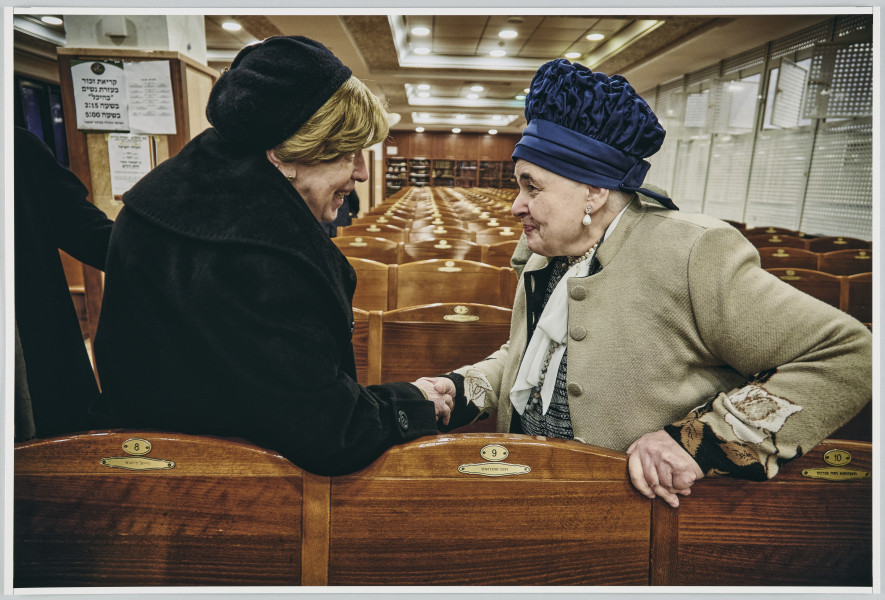 scena rodzajowa - Poziomy, barwny kadr fotograficzny przedstawiający dwie starsze kobiety ubrane w stroje wierzchnie oraz czapki, stojące na pierwszym planie na przeciwko siebie w dużym wnętrzu wypełnionym rzędami krzeseł, wydzielonym ażurową, białą ścianką z oknami. Kobiety witają się z serdecznością poprzez uścisk prawych dłoni.