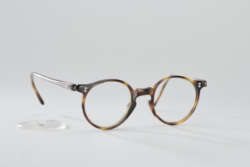 Okulary - Okulary czarno-brązowo-żółte (układ kolorów powtarzających się, naprzemiennie). Kształt szkieł zbliżony do koła. Na części oprawek od strony twarzy (front) - w rogach, w których łączona jest ta część z zausznikami, widoczne są - po dwa w każdym rogu - małe nity: płaskie, niewystające poza brzeg oprawki. Model okularów pozbawiony nanośników (tzw. nosków). Ślad po pęknięciu oprawki przy lewej tarczy, od strony części na nos. Przetarcia oprawki.