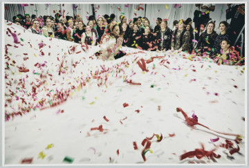 scena rodzajowa - Poziomy, barwny kadr fotograficzny przedstawiający scenę świętowania zaślubin. Większą część kadru wypełnia duża biała tkanina, na której widać dużo fragmentów kolorowych konfetti i serpentyn, które także spadają z góry. Nieostrość wskazuje na ruch tkaniny. Pośrodku tkaniny, w otworze, stoi kobieta w białym sukni stroju. Za tkaniną widoczna jest duża grupa dziewcząt z rozbawieniem przyglądająca się scenie. W tle – biała kotara oraz częściowo widoczne 2 sylwetki osób dokumentujących wydarzenie.
