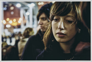 scena rodzajowa - Poziomy, barwny kadr fotograficzny przedstawiający zamyśloną młodą kobietę z długimi, opadającymi na ramiona włosami, w ciemnym stroju i z białym perłami na szyi. Za nią stoi inna kobieta, która patrzy w lewo, tło jest całkowicie nieostre, można wyodrębnić jedynie zarysy sylwetek ludzkich oraz punkty świetlne.