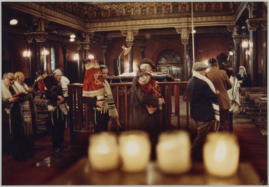 fotografia - Zdjęcie przedstawia grupę mężczyzn we wnętrzu Synagogi. Część z nich obchodzi bimę trzymając w rękach zwoje tory w ozdobnych sukienkach. Po lewej stronie kadru widać dwóch mężczyzn trzymających w rękach otwarte księgi. Modlą się. Wszyscy mężczyźni mają nakrycia głowy (jarmułki lub kaszkiety). Ramiona większości z nich okrywają tałesy. Po prawej stronie kadru, w głębi synagogi widać modlącą się kobietę w błękitnej chuście na głowie. U dołu kadru widoczne cztery palące się świece. Świece są rozmazane, ostrość jest skupiona na mężczyznach. 