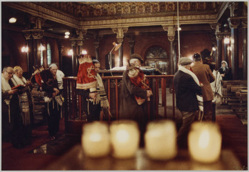fotografia - Zdjęcie przedstawia grupę mężczyzn we wnętrzu Synagogi. Część z nich obchodzi bimę trzymając w rękach zwoje tory w ozdobnych sukienkach. Po lewej stronie kadru widać dwóch mężczyzn trzymających w rękach otwarte księgi. Modlą się. Wszyscy mężczyźni mają nakrycia głowy (jarmułki lub kaszkiety). Ramiona większości z nich okrywają tałesy. Po prawej stronie kadru, w głębi synagogi widać modlącą się kobietę w błękitnej chuście na głowie. U dołu kadru widoczne cztery palące się świece. Świece są rozmazane, ostrość jest skupiona na mężczyznach. 