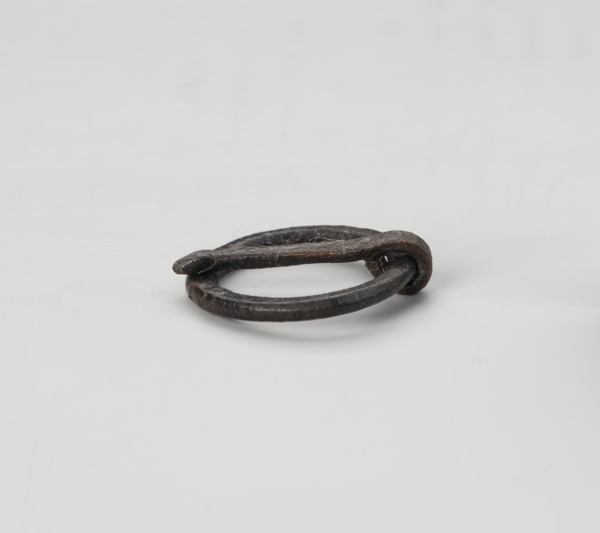 sprzączka do pasa - Ujęcie z tyłu; Kolista sprzączka żelazna ze spłaszczonego drutu z ruchomym kolcem na ramie. Kolec przymocowano poprzez wygięcie tępego końca drutu i owiniecie go wokół obręczy sprzączki. Na powierzchni zachowane są pojedyncze włókna.