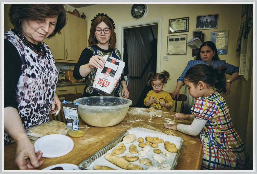 scena rodzajowa - Poziomy, barwny kadr fotograficzny przedstawiający kobietę i 4 dziewczynki w różnym wieku. Wszystkie zebrane są wokół stołu kuchennego, na którym widać proces przygotowywania ciast - stojące wokół stołu w pomieszczeniu (kuchni) i zajmujące się przygotowywaniem ciasta. Kobieta stoi po lewej stronie kadru, sięga po talerzyk (kadr ucina zawartość naczynia), obok niej stoi nastolatka w okularach wsypująca mąkę do miski, po prawej stronie kadru siedzą dwie małe dziewczynki zajęte lepieniem ciasta, z tyłu, za nimi stoi dziewczynka obserwująca pracę zebranych. Na stole widoczna jest forma z już uformowanymi ciastkami do pieczenia.