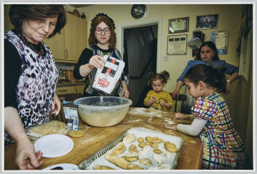 scena rodzajowa - Poziomy, barwny kadr fotograficzny przedstawiający kobietę i 4 dziewczynki w różnym wieku. Wszystkie zebrane są wokół stołu kuchennego, na którym widać proces przygotowywania ciast - stojące wokół stołu w pomieszczeniu (kuchni) i zajmujące się przygotowywaniem ciasta. Kobieta stoi po lewej stronie kadru, sięga po talerzyk (kadr ucina zawartość naczynia), obok niej stoi nastolatka w okularach wsypująca mąkę do miski, po prawej stronie kadru siedzą dwie małe dziewczynki zajęte lepieniem ciasta, z tyłu, za nimi stoi dziewczynka obserwująca pracę zebranych. Na stole widoczna jest forma z już uformowanymi ciastkami do pieczenia.