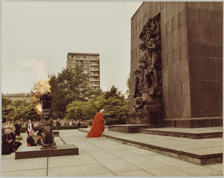 fotografia - Zdjęcie przedstawia uroczystości odbywające się przed Pomnikiem Bohaterów Getta w Warszawie. Na fotografii (po prawej stronie kompozycji) papież Jan Paweł II, ubrany w czerwoną pelerynę i białą piuskę kłania się bohaterom przedstawionym na centralnym panelu pomnika. Pomnik i uczestnicy wydarzenia widoczni są z boku (z profilu). Poniżej (po lewej stronie kadru) widać zapalone kamienne menory (stanowiące część instalacji miejsca pamięci). Poniżej widać kilka klęczących osób (w garniturach oraz strojach biskupich – różowych piuskach i pasach). 