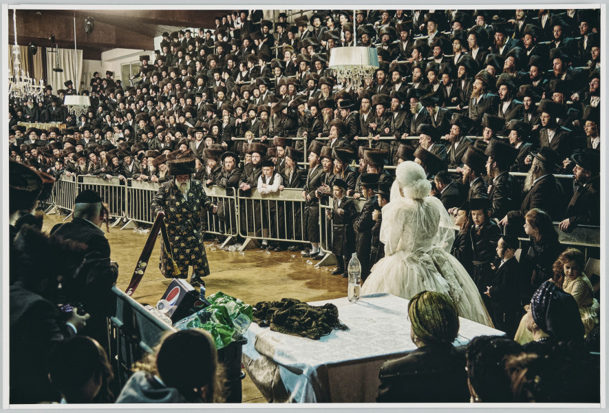 scena rodzajowa - Poziomy, barwny kadr fotograficzny, który w centralnej części przedstawia pannę młodą ubraną w strojną białą suknię ślubną, z białym czepcem na głowie i o twarzy zasłoniętej białym welonem oraz stojącego naprzeciwko niej starszego mężczyzny z siwą brodą w ozdobnym, stroju (dużą futrzaną, cylindryczną czapę, długi haftowany w złoty wzór płaszcz). Mężczyzna trzyma w rękach ozdobną, haftowaną szarfę. Oboje stoją po środku bardzo dużego pomieszczenia, w korytarzu utworzonym przed oddzielenie boków wnętrza przez metalowe barierki. Za barierkami, po obu stronach, utworzone są kaskadowo miejsca dla widzów (rodzaj stojących trybun). Tam stoi bardzo duża liczba mężczyzn z brodami i chłopców ubranych podobnie (duże futrzane, cylindryczne czapy – strajmły lub kapelusze, długie ciemne płaszcze – chałaty). Mężczyźni obserwują scenę rozgrywającą się poniżej, część z nich wygląda tak, jakby śpiewało. Po prawej stronie kadru, w rogu, za panną młodą stoi kwadratowy stół nakryty białym obrusem, na nim leży tkanina. Oddzielona tym stołem siedzi grupa kobiet w ciemnych strojach i z ozdobnymi czepkami na głowach. Widać także małą dziewczynkę.