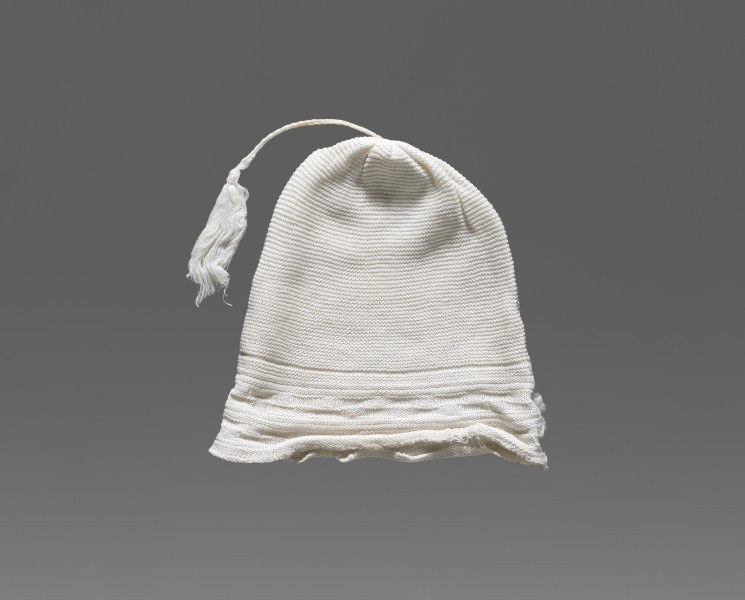 Czapeczka dziecięca w kolorze białym/écru. W dolnej części (około 1/3 wysokości od dolnej krawędzi) widoczny ozdobny geometryczny ścieg w formie szachownicy. W górnej części (po środku) przyszyty został sznurek zakończony frędzlem.