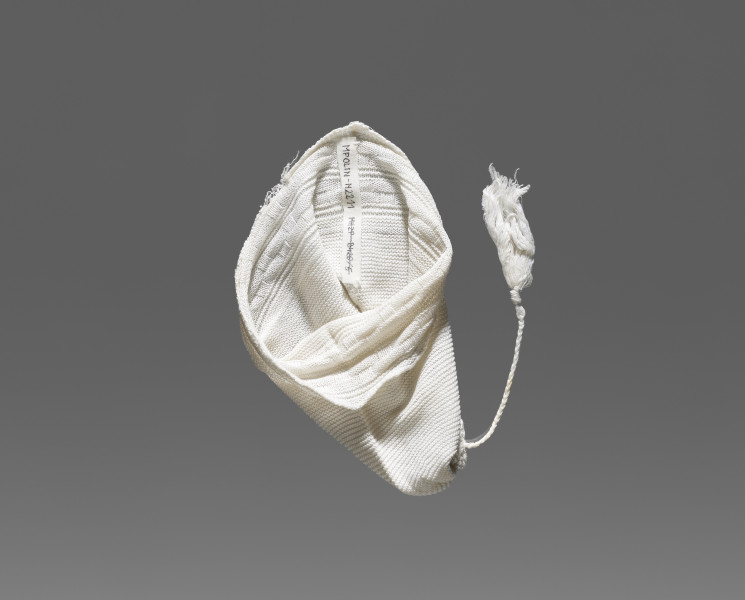 Czapeczka dziecięca w kolorze białym/écru. W dolnej części (około 1/3 wysokości od dolnej krawędzi) widoczny ozdobny geometryczny ścieg w formie szachownicy. W górnej części (po środku) przyszyty został sznurek zakończony frędzlem.