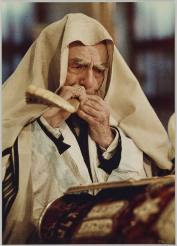 fotografia - Zdjęcie przedstawia mężczyznę stojącego przed pulpitem i dmącego w róg (Szofar - róg wykorzystywany podczas obchodów święta Rosz ha-Szana). Głowa mężczyzny jest okryta szalem modlitewnym (tałesem). Spod tałesu wystają białe płócienne rękawy oraz mankiety garnitury i białej koszuli wystające spod spodu.