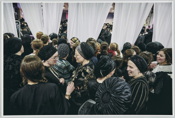 scena rodzajowa - Na pierwszym planie poziomego, barwnego kadru fotograficznego widać dużą grupę, stojących, rozmawiających ze sobą kobiet. Cześć z nich patrzy za białe zasłony, przedzielające pomieszczenie, w którym są na dwie części. Kobiety są ubrane w czarne, stroje, częściowo z elementami dekoracyjnymi lub wzorami w innych kolorach. Niektóre z nich na głowach noszą czepki. Za zasłonami, które wiszą od sufitu w kilku częściach, między którymi są prześwity, widać w bardzo ograniczonym stopniu dalszą część pomieszczenia wypełnioną tłumem mężczyzn w czarnych strojach, kapeluszach lub okrągłych nakryciach głowy (jarmułkach).