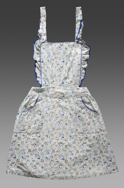 Fartuszek dziecięcy wykonany z tkaniny w kolorze écru z ozdobnym kolorowym nadrukiem – drobne wizerunki kwiatów, liści, motyli, ważek i grzybów (muchomorów). Nadruk w kolorach: zielonym, niebieskim i pomarańczowym. Fartuch składa się z prostokątnego fragmentu materiału obszytego po bokach falbanką wykończoną niebieską lamówką (karczek/część piersiowa), paska oraz spódnicy. Karczek, pasek oraz spódnica zostały ze sobą zszyte. Do górnych krawędzi karczka przyszyte zostały długie pasy tkaniny (umożliwiające jego zawiązanie na szyi). Pasek zakończony jest na obu końcach pętelkami wykonanymi z tej samej tkaniny (miejsce na guzik lub tasiemkę). Do spódnicy zostały przyszyte kieszenie obszyte dodatkowo ozdobną niebieską lamówką.