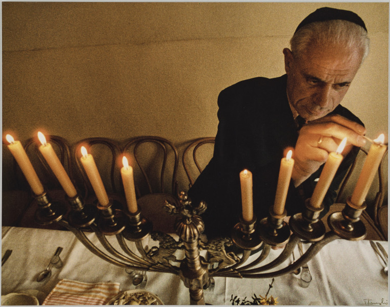 fotografia - Zdjęcie przedstawia mężczyznę zapalającego świece u umieszczone w kandelabrze stojącym na przykrytym białym obrusem stole. Świecznik ma osiem ramion - po cztery na każdą ze stron trzonu. Trzon zwieńczony jest ozdobnym orłem w koronie. Wszystkie osiem świec już płonie – mężczyzna na zdjęciu właśnie podpalił ostatnią z nich. Na fragmencie stołu, który został ujęty w kadrze widać sztućce oraz zastawę– został nakryty. Mężczyzna ma na sobie garnitur oraz jarmułkę. Pod ścianą widać rząd krzeseł. 