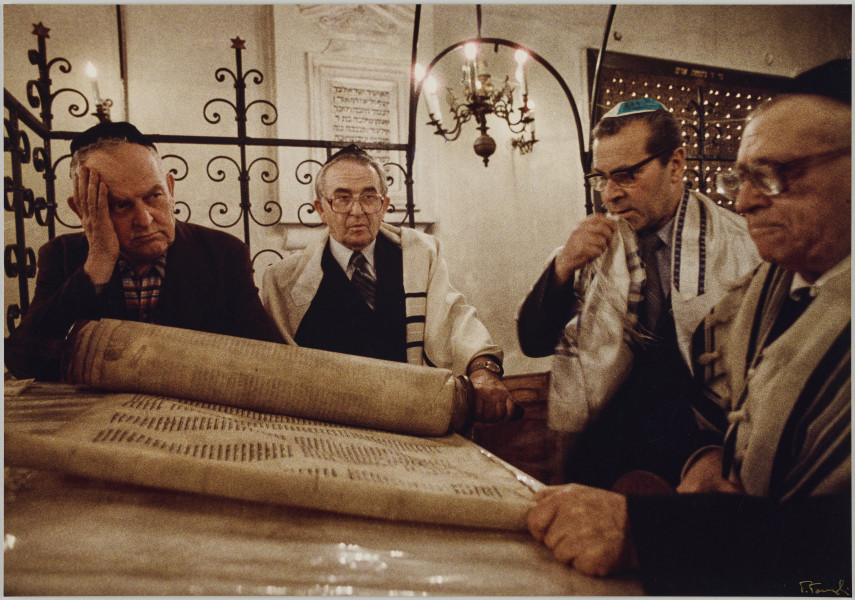 fotografia - Zdjęcie przedstawia czterech mężczyzn pochylających się nad rozłożonym na pulpicie zwojem Tory. Wszyscy mają na głowach jarmułki. Ramiona trzech z nich (kolejno od prawej strony) są okryte tałesem. Ci mężczyźni mają również na sobie okulary. Za nimi widać kratowanie oddzielające przestrzeń bimy od pozostałych przestrzeni Synagogi. W tle widoczny także ozdobny żyrandol i tablica pamiątkowa.