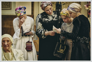 scena rodzajowa - Poziomy, barwny kadr fotograficzny przedstawiający grupę 5 kobiet we wnętrzu. W lewym dolnym rogu kompozycji znajduje się starsza kobieta (widać jej popiersie) w jasnym stroju i jasnymi koralami na szyi oraz z jasną chusta na głowie. Za nią stoją 4 dalsze kobiety – jedna w białej sukience, białych perłach na szyi i kolorowej chustce na głowie czyta z książeczki, którą trzyma w lewej ręce. Prawą ręką przysłania sobie oczy w geście skupienia. Pozostałe 3 kobiety stoją obok siebie. Ubrane są w ciemne suknie i kolorowe chusty na głowach, na szyi mają sznurki białych pereł. Kobiety patrzą w tekst, który trzyma jedna z nich.Inne fotografie prezentujące ten ślub: MPOLIN-M1539 i MPOLIN-M1541.