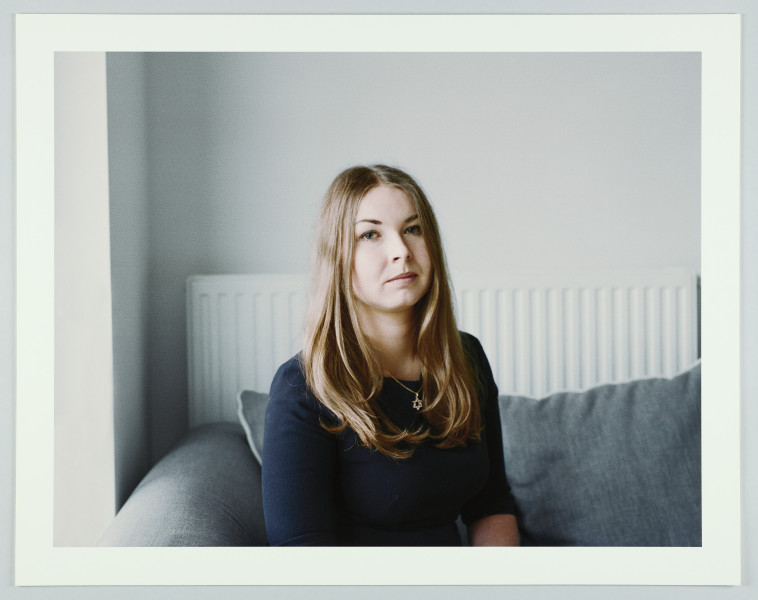 portret fotograficzny - Fotografia barwna skadrowana poziomo przedstawiająca półpostaciowy portret kobiety. Portretowana ma długie, proste jasne włosy do ramion uczesanych z przedziałkiem na środku głowy i puszczonych luźno na ramiona, ubrana jest w szaroniebieską bluzkę z długim rękawem podwiniętym pod łokcie, patrzy się bezpośrednio w stronę widza. Kobieta siedzi (o czym świadczy ułożenie rąk) we wnętrzu na tle białej ściany, na szarej sofie, której oparcie widać w tle. Na ścianie, za kanapą, widoczny jest duży kaloryfer płytowy. Z boku, po lewej stronie widoczny jest fragment wnęki okiennej.