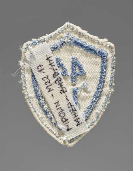 Tarcza szkolna w formie naszywki wykonana z tkaniny w kolorze białym. Czworoboczna - z trzema rogami na górze i jednym na dole, każda krawędź zaokrąglona: górne do wewnątrz (do dołu), boczne na zewnątrz. Krawędzie obszyte nicią w kolorze niebieskim. W centralnym polu wszyty maszynowo nicią w tym samym kolorze numer ,,44’’ (nr szkoły) oraz literę ,,L'' pod spodem.