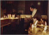 fotografia - Zdjęcie przedstawia mężczyznę podczas modlitwy wśród zapalonych świec. Mężczyzna znajduje się w głębi niewielkiego pomieszczenia. Przed nim stoją stoły, a na nich duże naczynia, w których ustawione są zapalone świece. Mężczyzna ma na sobie garnitur oraz jarmułkę. Jego ramiona są okryte tałesem. Trzyma w rękach księgę. Jest lekko pochylony w stronę tekstu. 