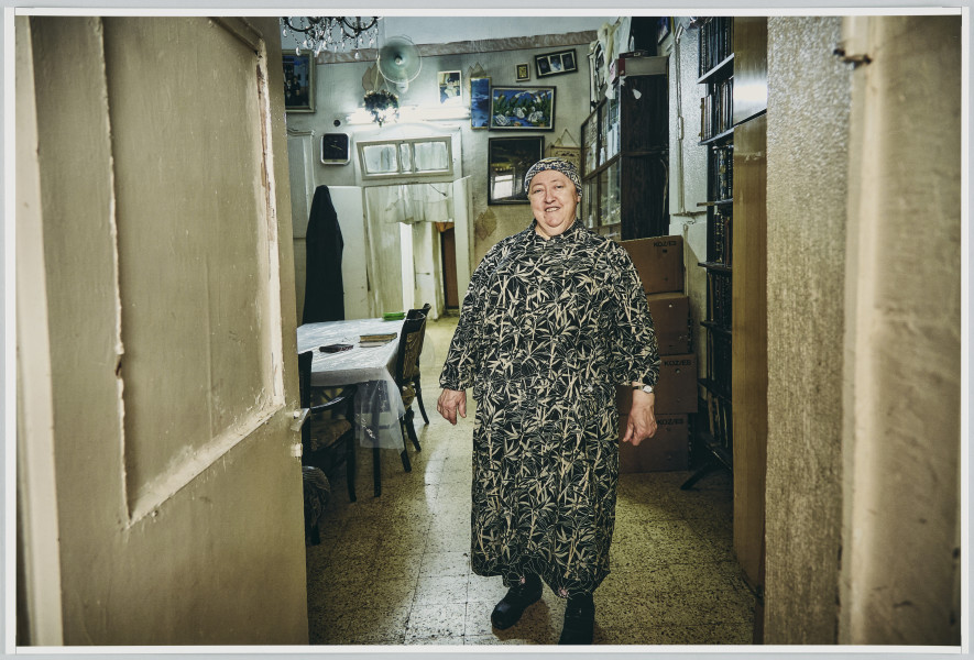 scena rodzajowa - Poziomy, barwny kadr fotograficzny przedstawiający całopostaciowy portret dojrzałej, korpulentnej kobiety ubranej w czarno-białą długą sukienkę o kwiatowym wzorze oraz chustę na głowie. Kobieta stoi u progu pokoju – przez otwarte drzwi widoczne po lewej stronie. We wnętrzu widać po prawej stronie regały z książkami oraz kartonowe pudła stojące na podłodze, po lewej – stół przykryty obrusem i folią oraz 3 tapicerowane krzesła. Na suficie wisi ozdobny żyrandol (widoczny fragment). W głębi widoczna ściana, na której wisi lustro, wentylator elektryczny, obrazi oraz zdjęcia w ramach. W ścianie znajduje się futryna z otwartymi, dwuskrzydłowymi drzwiami prowadzącymi do dalszego pomieszczenia. Na lewym ich skrzydle wisi czarny, długi ubiór (płaszcz?).