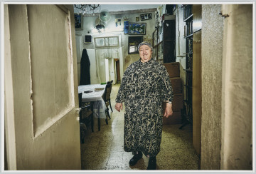 scena rodzajowa - Poziomy, barwny kadr fotograficzny przedstawiający całopostaciowy portret dojrzałej, korpulentnej kobiety ubranej w czarno-białą długą sukienkę o kwiatowym wzorze oraz chustę na głowie. Kobieta stoi u progu pokoju – przez otwarte drzwi widoczne po lewej stronie. We wnętrzu widać po prawej stronie regały z książkami oraz kartonowe pudła stojące na podłodze, po lewej – stół przykryty obrusem i folią oraz 3 tapicerowane krzesła. Na suficie wisi ozdobny żyrandol (widoczny fragment). W głębi widoczna ściana, na której wisi lustro, wentylator elektryczny, obrazi oraz zdjęcia w ramach. W ścianie znajduje się futryna z otwartymi, dwuskrzydłowymi drzwiami prowadzącymi do dalszego pomieszczenia. Na lewym ich skrzydle wisi czarny, długi ubiór (płaszcz?).