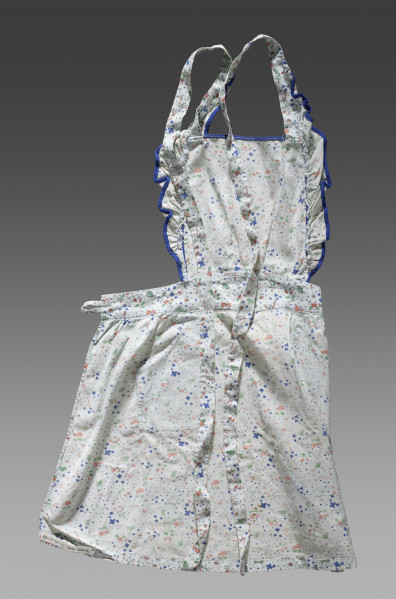 Fartuszek dziecięcy wykonany z tkaniny w kolorze écru z ozdobnym kolorowym nadrukiem – drobne wizerunki kwiatów, liści, motyli, ważek i grzybów (muchomorów). Nadruk w kolorach: zielonym, niebieskim i pomarańczowym. Fartuch składa się z prostokątnego fragmentu materiału obszytego po bokach falbanką wykończoną niebieską lamówką (karczek/część piersiowa), paska oraz spódnicy. Karczek, pasek oraz spódnica zostały ze sobą zszyte. Do górnych krawędzi karczka przyszyte zostały długie pasy tkaniny (umożliwiające jego zawiązanie na szyi). Pasek zakończony jest na obu końcach pętelkami wykonanymi z tej samej tkaniny (miejsce na guzik lub tasiemkę). Do spódnicy zostały przyszyte kieszenie obszyte dodatkowo ozdobną niebieską lamówką.