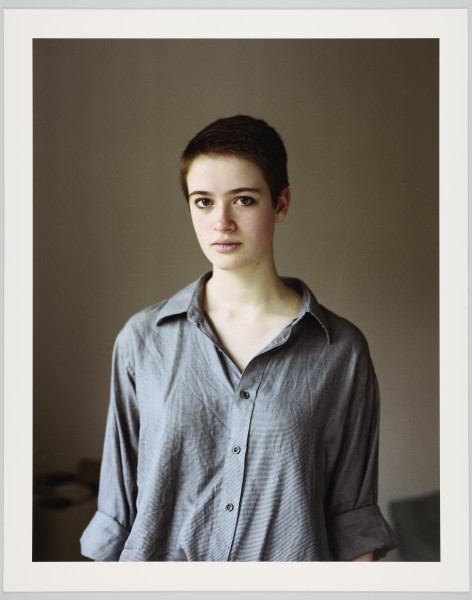 portret fotograficzny - Fotografia barwna skadrowana pionowo przedstawiająca półpostaciowy portret młodej kobiety. Portretowana dziewczyna o ciemnych, bardzo krótkich włosach, ubrana w szarą koszulę zapinaną na guziki, o szeroko rozpiętym kołnierzyku, z długimi rękawami podwiniętymi do wysokości łokcia, patrzy się bezpośrednio w stronę widza. Kobieta stoi we wnętrzu na tle jednobarwnej, szarej ściany.