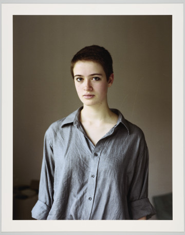 portret fotograficzny - Fotografia barwna skadrowana pionowo przedstawiająca półpostaciowy portret młodej kobiety. Portretowana dziewczyna o ciemnych, bardzo krótkich włosach, ubrana w szarą koszulę zapinaną na guziki, o szeroko rozpiętym kołnierzyku, z długimi rękawami podwiniętymi do wysokości łokcia, patrzy się bezpośrednio w stronę widza. Kobieta stoi we wnętrzu na tle jednobarwnej, szarej ściany.