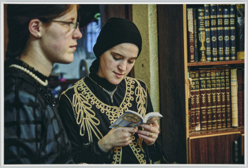 scena rodzajowa - Poziomy, barwny kadr fotograficzny przedstawiający dwie młode kobiety stojące we wnętrzu. Sylwetka jednej z nich, w okularach, w czarnej bluzce i krótkim sznurem białych pereł na szyi, widoczna z prawego profilu, wypełnia lewą stronę kadru. Dziewczyna patrzy przed siebie. Druga kobieta stoi koło niej. Jest ubrana w czarna bluzkę, czarną kamizelkę ozdobioną złotą wstążką w duże wzory, nosi na szyi srebrny niewielki naszyjnik oraz czarną chustę na głowie. W ręku trzyma książeczkę w języku hebrajskim i czyta z niej. Po prawej stronie widoczny jest ciemnobrązowy regał wypełniony ściśle książkami o ozdobnych grzbietach.