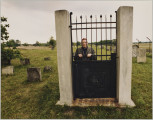 fotografia - Zdjęcie przedstawia mężczyznę stojącego za zamkniętą bramą. Brama składa się z gładkiej metalowej płyty sięgającej połowy wysokości, pionowych prętów o ozdobnych zwieńczeniach (od połowy wysokości do góry) oraz prętów poziomych (dwóch na górze i jednego na dole). Mężczyzna opiera się rękami o jeden z poziomych prętów bramy (umieszczony najbliżej płyty). Patrzy przed siebie. Ma na sobie ciemną kurtkę oraz jarmułkę. Brama jest umocowana pomiędzy dwoma betonowymi słupami. Brakuje jednak reszty ogrodzenia. Za bramą widoczna łąka oraz wyrastające z ziemi pojedyncze macewy i drzewa. 