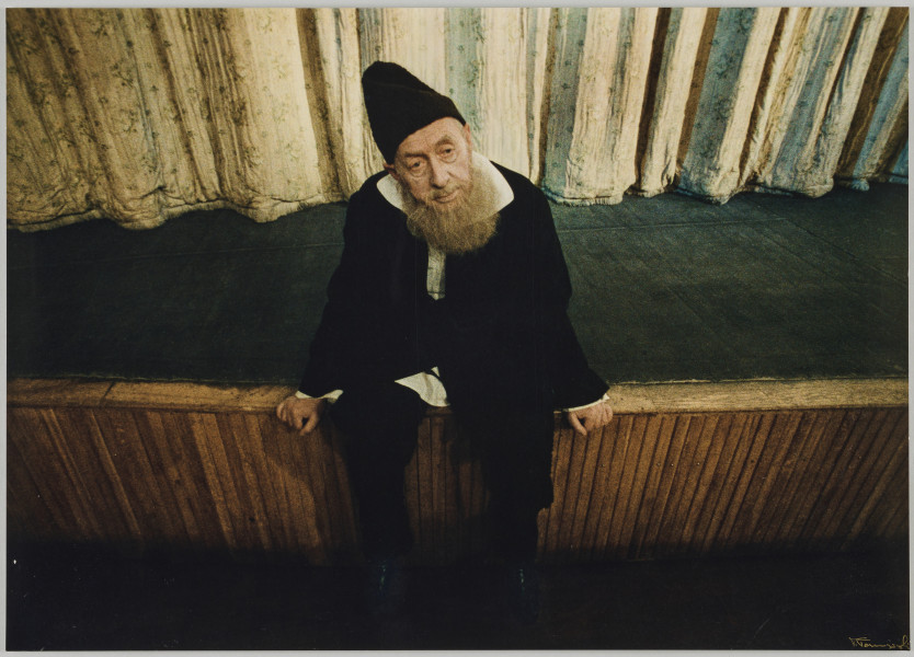 fotografia - Zdjęcie przedstawia starszego mężczyznę, aktora z siwą brodą siedzącego na krawędzi sceny teatralnej. Mężczyzna ma na sobie czarne spodnie, białą koszulę i czarne okrycie wierzchnie (chałat?). Mężczyzna opiera się rękami o krawędź podestu. Nogi ma spuszczone luźno do dołu. Za nim widać fragment jasnej kurtyny.