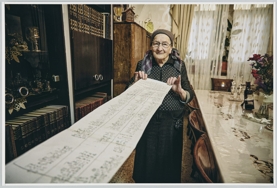 scena rodzajowa - W centralnej części poziomego, barwnego kadru stoi starsza kobieta w okularach ubrana w czarną sukienkę z górą w białe kropki, na głowie ma chustkę. Poziomo, w perspektywie pod skosem, od strony lewego dolnego rogu kadru, rozciąga się długi zwój papieru którego koniec kobieta trzyma w rękach. Na zwoju widoczne są odręczne zapiski wykonane na czarno. Kobieta znajduje się we wnętrzu pokoju. Po lewej stronie przy ścianie stoją regały z książkami i bibelotami oraz drewniana szafa, zaś po prawej – długi stół, którego widać fragment, z 4 krzesłami. Na stole leży biały, haftowany obrus przykryty przezroczystą folią, wazon z kwiatami, butelka wina w stojaku oraz 2 srebrne świeczniki. W głębi znajduje się okno, na parapecie którego stoją kwiaty doniczkowe, zasłonięte białą, gęstą, dwuczęściową firanką, po obu jego bokach wiszą, dekoracyjnie upięte, beżowe zasłony.