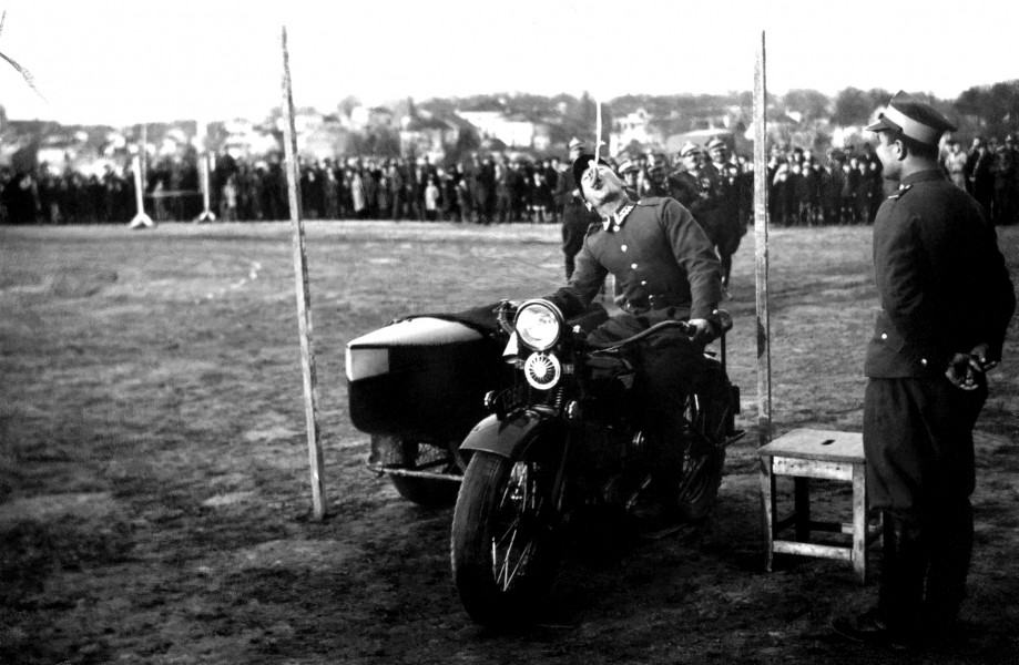 Zdjęcie ukazuje zawody w wyszkoleniu indywidualnym w jeździe na motocyklu. Mężczyzna na motocyklu przejeżdzający pod przeszkodą, z otwartymi ustami próbując chwycić wiszący przedmiot.