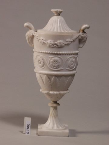 Wazon w stylu klasycystycznym z uchwytem w kształcie głów baranów. Pokrywka dzwonowata.